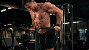 Muscular man shirtless, sweating, performing a dip exercise on parallel bars in a dim gym.